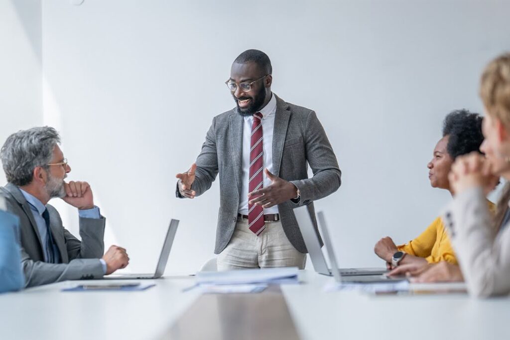 A man in a suit stands and speaks confidently to four colleagues seated at a conference table with laptops open.