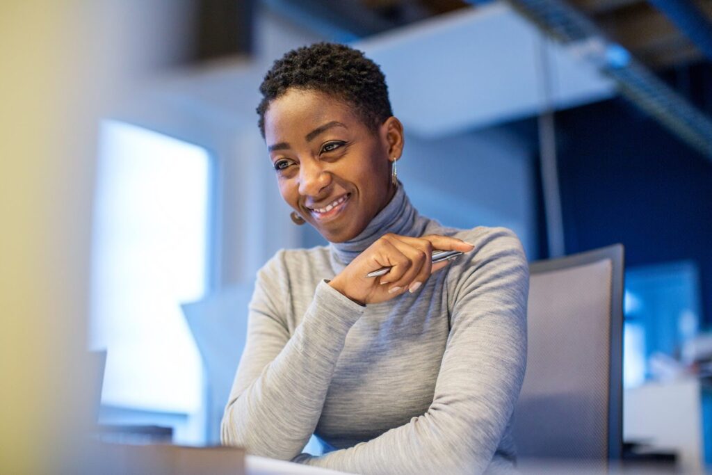 A woman in business attire seated in a modern office space smiles at her desk.