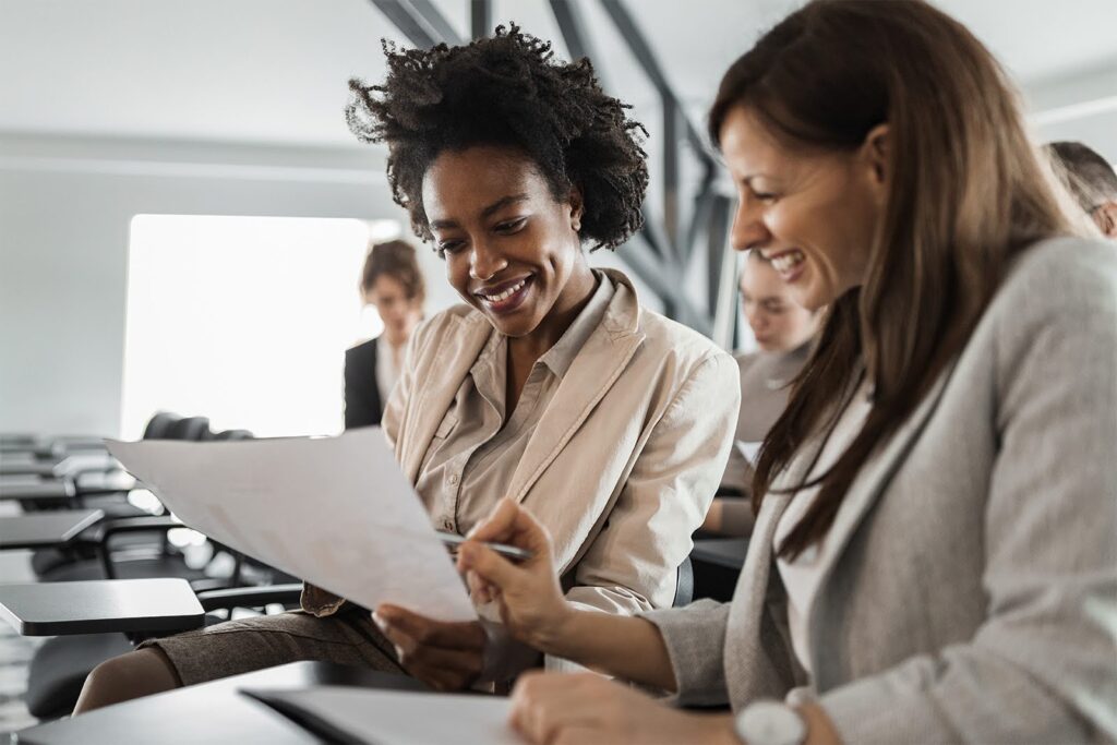 Two smiling women review documents together in a bright, modern office setting.