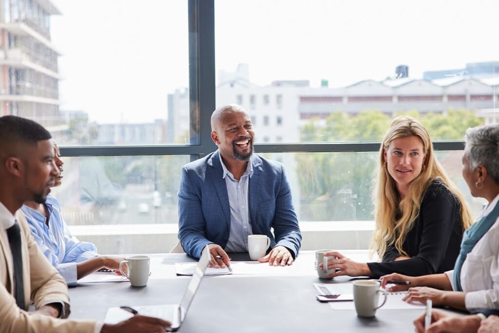A diverse group of professionals smiles and converses around a conference table in a conference room with large windows.