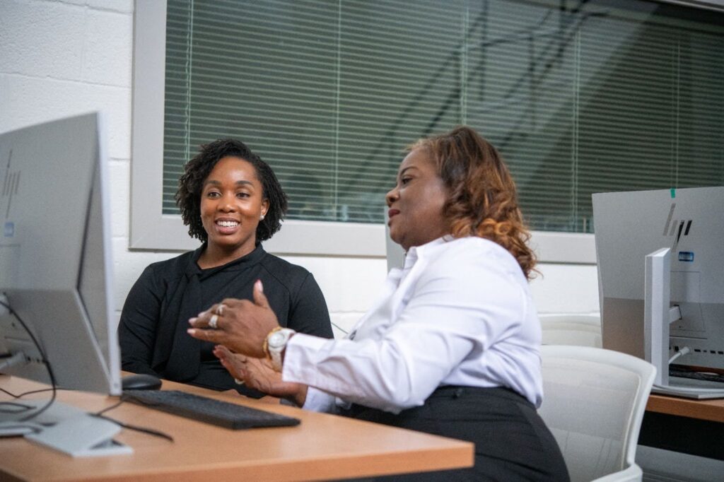 Two students converse as they work together at a desktop computer.