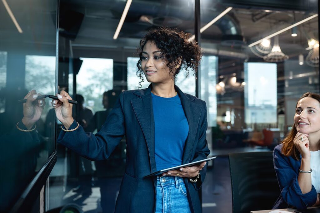 In a modern office setting, a woman holding a tablet writes on a glass board during a meeting while a colleague seated nearby listens and smiles.