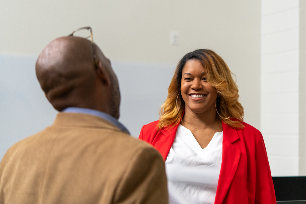 A smiling Black woman in a red blazer and white blouse makes eye contact with a man in a brown blazer during a professional conversation.