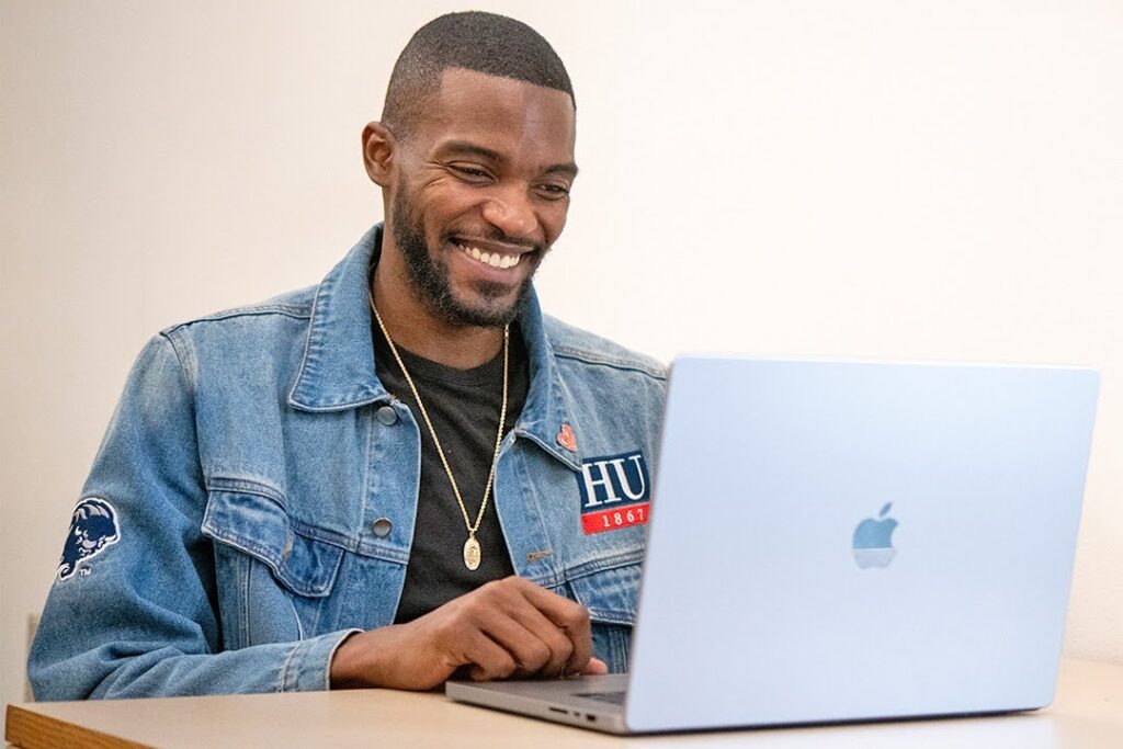A smiling man wearing a jean jacket with Howard University branding uses a laptop computer.