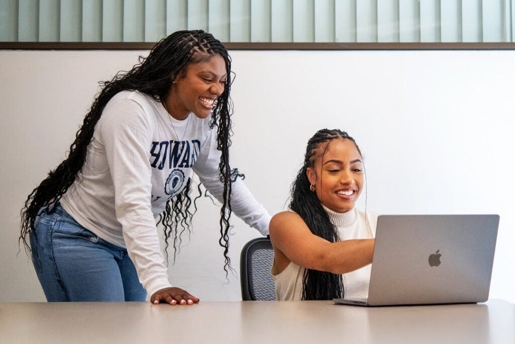 Two young women laugh while looking at a laptop screen together.